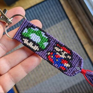 Könnte beinhalten: Ein handgefertigter, lila Schlüsselanhänger mit pixeligen Bildern eines grünen Pilzes und Mario. Der Schlüsselanhänger hat einen silbernen Verschluss und eine lila Quaste mit roten Akzenten. Das Design erinnert an klassische Videospiele.