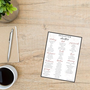 Puede incluir: Una lista de verificación de limpieza en papel blanco con borde negro, que enumera categorías como ropa, electrónica y cocina. El papel está sobre una superficie de madera, con una taza de café, un bolígrafo y un bloc de notas.