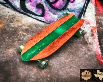 Könnte beinhalten: Ein Skateboard aus Holz mit einer grünen Epoxidharz-Einlage. Das Deck ist aus rotem Holz und hat eine abgerundete Nase und ein abgerundetes Heck. Das Skateboard hat schwarze Räder und Achsen.