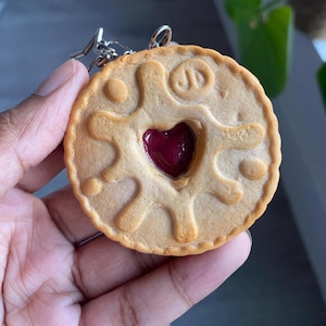 May include: A round, tan-coloured biscuit-shaped keyring featuring a scalloped edge and a central heart-shaped cutout filled with a dark red, translucent jam-like material. The surface has embossed circular patterns and the letters JD are visible.
