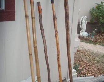 May include: A collection of handcrafted walking sticks and canes, leaning against a wall. The sticks vary in color and material, including bamboo and natural wood. Some have decorative elements like wrapped wire or unique carved handles. The image also shows two decorative stones with text.