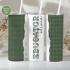May include: Three stainless steel tumblers with white straws. Two tumblers feature a green and black checkered pattern. The center tumbler has the words "Educator Nurse Olivia" in green and black. The tumblers are designed for beverages.