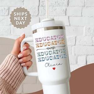 May include: White tumbler with a handle and a straw. The tumbler has the words "EDUCATOR" and "nurse" in colorful text. Below the text is the name "Olivia" with a red heart. The tumbler is held by a person.