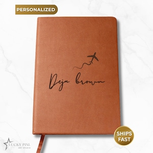 May include: A rectangular notebook with a textured tan cover. The front features a black graphic of an airplane with a trailing flight path line above the black cursive text that reads Deja brown. A small fabric bookmark extends from the bottom.
