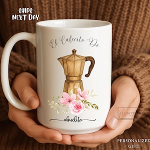 May include: White ceramic mug featuring a gold-colored coffee maker illustration, pink flowers, and the text "El Cafecito De abuelito." The mug has a large handle and is held by a person.
