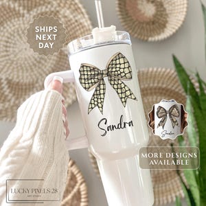 May include: White tumbler with a bow design and the name "Sandra" in black script. The bow has a snakeskin pattern. The tumbler has a handle and a straw. The text "More designs available" is also visible.