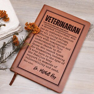 May include: A rectangular brown notebook with a black border and printed text that defines a veterinarian as an authentic individual with passion for all living beings. The cover includes the name Dr. Mitchell Ray and is decorated with orange floral sprigs.