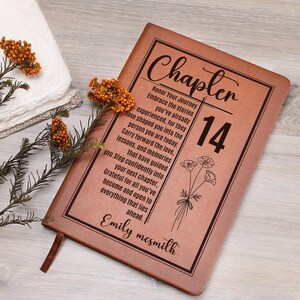 May include: A tan leather-like journal featuring the text Chapter 14, a floral illustration, and a quote about life journeys. The cover is personalized with the name Emily Mcsmith in script font, enclosed within a black rectangular border.