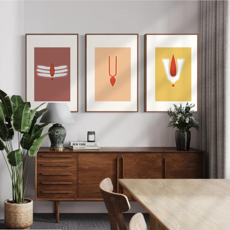 May include: Three framed prints with minimalist designs featuring Hindu religious symbols in red, white, and orange on a peach, yellow, and brown background. The prints are hanging on a white wall above a wooden dresser.