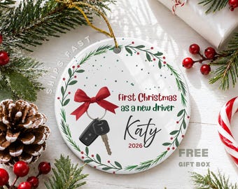 May include: A round white ceramic ornament featuring a green pine wreath with red berries. The center displays a black car key fob with a red bow, the text First Christmas as a new driver, the name Katy, and the year 2026 in black and red script.
