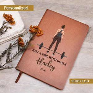 May include: A brown leather-textured notebook featuring a graphic of a person standing behind a barbell. The text reads JUST A GIRL WITH GOALS, the name Hailey, and the year 2026. The cover has a smooth finish and a rectangular shape.