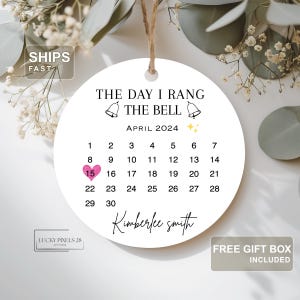 May include: A white, round ornament with the text "THE DAY I RANG THE BELL" and a calendar for April 2024, highlighting the 15th. Includes the name "Kimberlee smith" and a jute string for hanging.