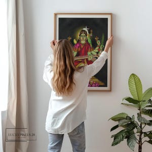 May include: A framed print of a colorful painting depicting a Hindu goddess playing a stringed instrument. The goddess is depicted with multiple arms and is adorned with jewelry and flowers. The background is dark and features a crescent moon.