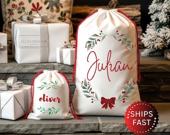 May include: White drawstring Christmas gift bags with red accents. One bag has the name "Julian" in red script, surrounded by a green and red wreath. A smaller bag has the name "Oliver". Presents wrapped in white paper are in the background.