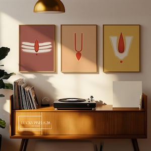 May include: Three framed prints with minimalist designs of Hindu religious symbols in red, white, and gold on a peach, beige, and yellow background. The prints are hanging on a white wall above a wooden cabinet with a record player on top.