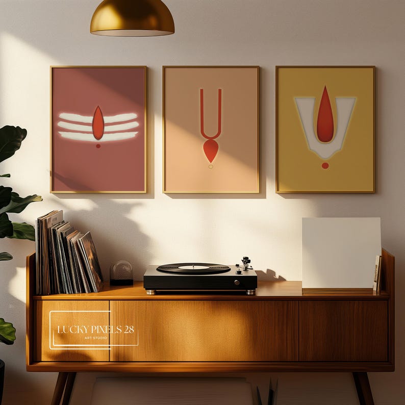 May include: Three framed prints with minimalist designs of Hindu religious symbols in red, white, and gold on a peach, beige, and yellow background. The prints are hanging on a white wall above a wooden cabinet with a record player on top.