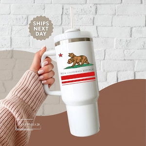 May include: White travel mug with a handle, featuring the New California Republic flag design. The flag includes a brown bear, a red star, and red stripes. The mug has a white lid and a straw.