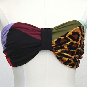 May include: A colorful bandeau top with a bow design. The top features a patchwork of colors including black, purple, red, olive green, and a leopard print pattern. The bow is centered with a black band.
