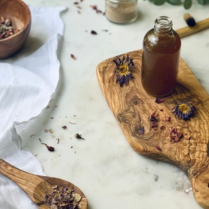 May include: A clear glass bottle with a brown liquid inside, sitting on a wooden cutting board with dried flowers. The bottle is on a white marble surface with a white cloth and a small bowl of dried flowers.