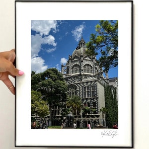 May include: A framed print of a building with a patterned facade and a domed roof, set against a blue sky with clouds. Lush green trees surround the building. The print is held by a hand with pink nail polish.