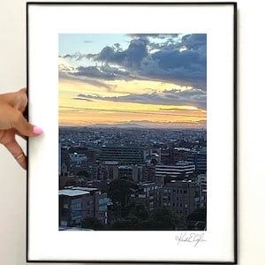 May include: A framed photograph of a cityscape at sunset. The sky is filled with dramatic clouds, transitioning from blue to orange and yellow hues. The city buildings are silhouetted against the colorful sky. The photograph is held by a person.