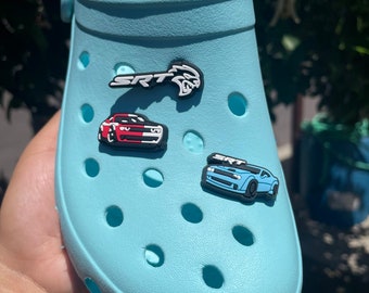 May include: Light blue clogs with three car-themed shoe charms. The charms are a red and white car with the word "SRT" on it, a black and white SRT logo, and a blue car with the word "SRT" on it.