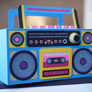 May include: A colorful, retro-style boombox with a blue body, yellow accents, and pink and black details. The design includes speakers, a cassette tape, and a handle. The overall aesthetic is reminiscent of the 1980s.
