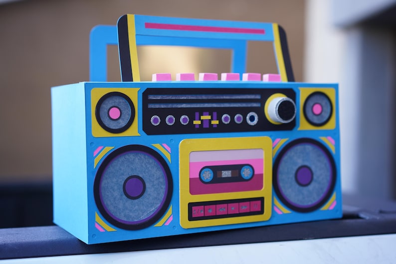 May include: A colorful, retro-style boombox with a blue body, yellow accents, and pink and black details. The design includes speakers, a cassette tape, and a handle. The overall aesthetic is reminiscent of the 1980s.