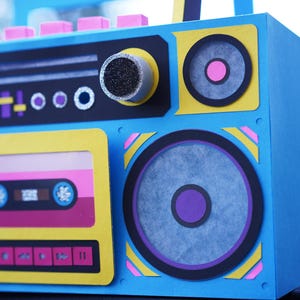 May include: A blue, yellow, and pink boombox with a cassette tape design. The speakers have purple and pink accents. The boombox has a retro aesthetic, with a focus on the 1980s and 1990s.