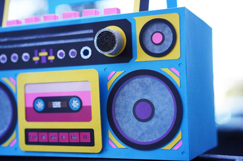 May include: A blue, yellow, and pink boombox with a cassette tape design. The speakers have purple and pink accents. The boombox has a retro aesthetic, with a focus on the 1980s and 1990s.