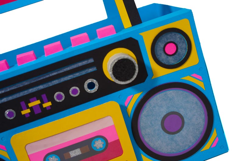 May include: A colorful, retro-style boombox with a vibrant design. The boombox features a blue body with yellow and black accents, pink details, and a cassette tape. The speakers have purple and pink centers.