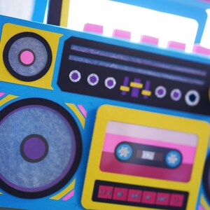 May include: A colorful, retro-style boombox in shades of blue, yellow, pink, and black. The design includes speakers, a cassette tape deck, and various control buttons. The boombox has a handle and is decorated with geometric shapes.