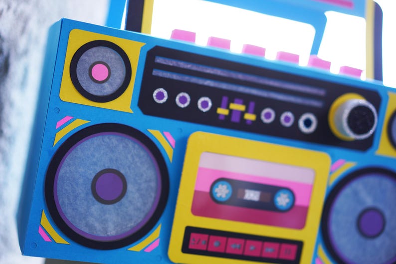 May include: A colorful, retro-style boombox in shades of blue, yellow, pink, and black. The design includes speakers, a cassette tape deck, and various control buttons. The boombox has a handle and is decorated with geometric shapes.