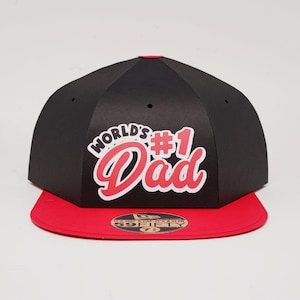 Puede incluir: Gorra de béisbol negra y roja con el texto "WORLD'S #1 Dad" en blanco y rojo. La gorra tiene una visera plana roja y una corona negra. Un accesorio de moda para padres.