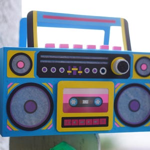 May include: A colorful, retro-style boombox with a blue body, yellow accents, and pink and purple details. The design includes speakers, a cassette tape, and a handle. The boombox is a vibrant piece of art.