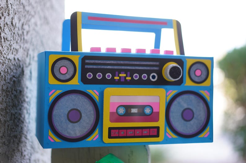 May include: A colorful, retro-style boombox with a blue body, yellow accents, and pink and purple details. The design includes speakers, a cassette tape, and a handle. The boombox is a vibrant piece of art.