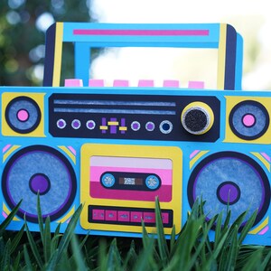 May include: A colorful, handmade boombox with a retro design. The blue, yellow, and pink accents create a vibrant aesthetic. Features include speaker details, a cassette tape, and a handle. The boombox is set against a backdrop of green grass.