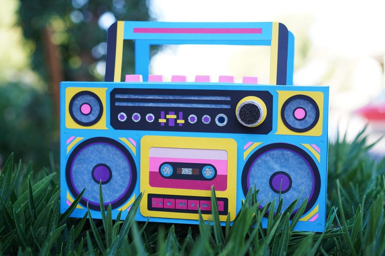 May include: A colorful, handmade boombox with a retro design. The blue, yellow, and pink accents create a vibrant aesthetic. Features include speaker details, a cassette tape, and a handle. The boombox is set against a backdrop of green grass.