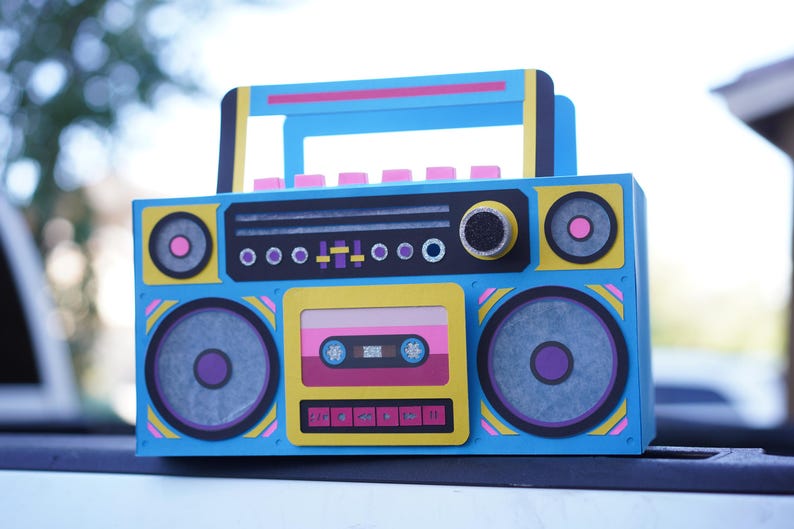 May include: A colorful, retro-style boombox with a blue body, yellow accents, and pink and purple details. The design includes speakers, a cassette tape, and a handle, evoking an 80s aesthetic. The boombox is a decorative item.