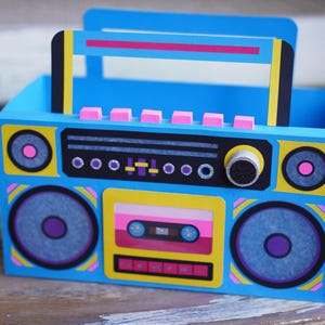 May include: A blue, yellow, and pink boombox-shaped box with a handle. The box features speaker graphics, a cassette tape design, and a control panel. The design is reminiscent of 1980s retro style.