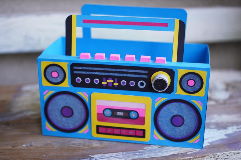 May include: A blue, yellow, and pink boombox-shaped box with a handle. The box features speaker graphics, a cassette tape design, and a control panel. The design is reminiscent of 1980s retro style.