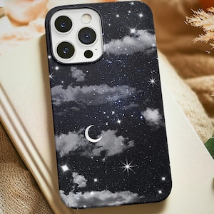 Puede incluir: Una funda negra para smartphone con un diseño de cielo nocturno que presenta una luna creciente, estrellas y nubes. La funda está sobre un libro cerrado con una cubierta de color crema. El fondo incluye una tela suave y texturizada.
