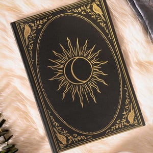 May include: Black journal with a gold sun and moon design on the cover. The cover is framed with a gold border and floral and bird accents. The journal is lying on a fluffy, white surface.