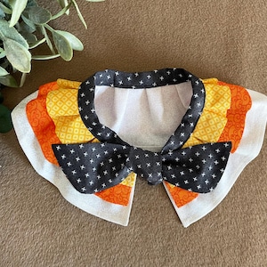 May include: A white, black, orange, and yellow fabric collar with a black bow tie. The collar has a ruffled edge and is decorated with a geometric pattern.