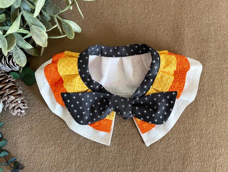 May include: A white, black, orange, and yellow fabric collar with a black bow tie. The collar has a ruffled edge and is decorated with a geometric pattern.