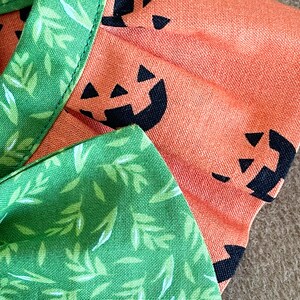 May include: A green fabric with white leaf designs and a bow. The fabric is sewn to a peach fabric with black pumpkin faces.
