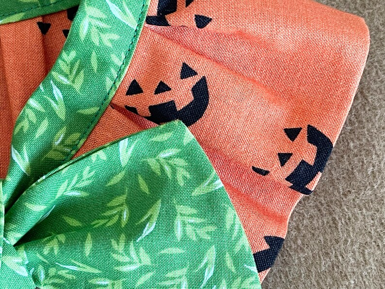 May include: A green fabric with white leaf designs and a bow. The fabric is sewn to a peach fabric with black pumpkin faces.