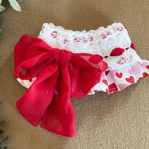 May include: A white and red heart print fabric with a white lace trim and a large red bow. The fabric is gathered and ruffled.