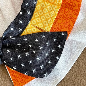 May include: A black fabric bow with white plus signs on it. The bow is attached to a white fabric with a yellow and orange fabric underneath.