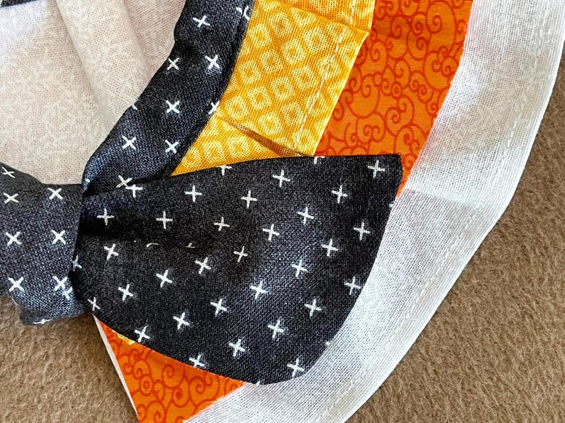 May include: A black fabric bow with white plus signs on it. The bow is attached to a white fabric with a yellow and orange fabric underneath.